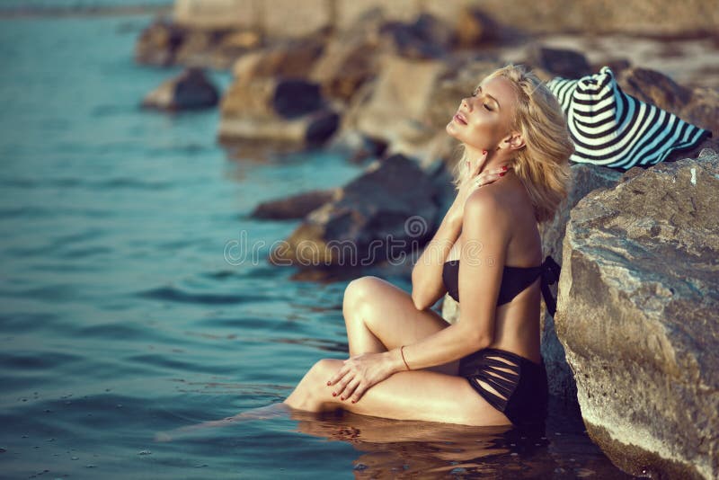 Blonde atractivo bronceado magnífico en el traje de baño negro que se sienta en el agua en las piedras grandes que acaricia su cu imagen de archivo libre de regalías Blonde atractivo bronceado magnífico en el traje de baño negro que se sienta en el agua en las piedras grandes que acaricia su cu imagen de archivo libre de regalías