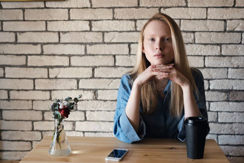 Blonde in blue shirt sits at the table in a cafe on which stands a black paper cup with coffee and looks at the camera stock photo Blonde in blue shirt sits at the table in a cafe on which stands a black paper cup with coffee and looks at the camera stock photo