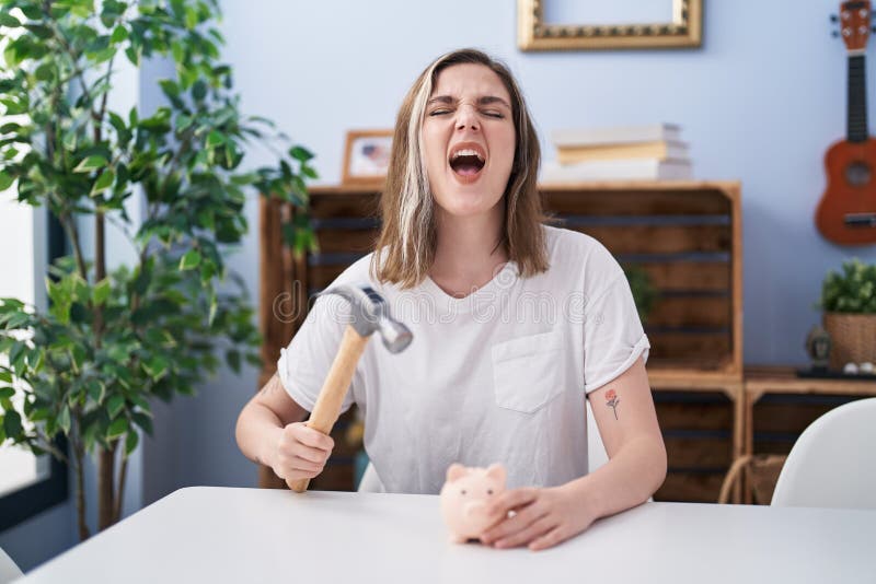 Blonde caucasian woman holding piggy bank and hammer angry and mad screaming frustrated and furious, shouting with anger looking