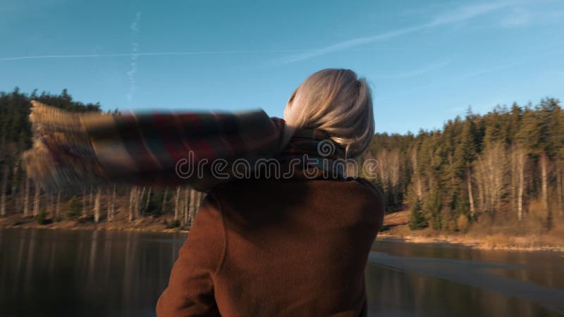 Blonde in a coat and scarf stands by the lake