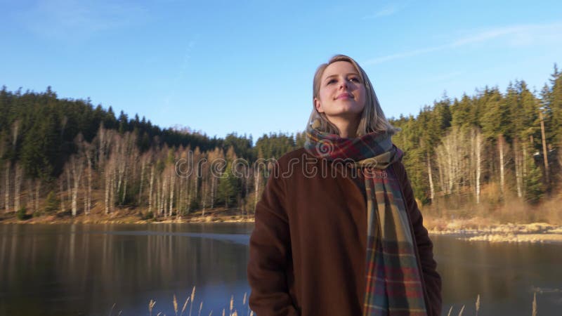 Blonde in a coat and scarf stands by the lake