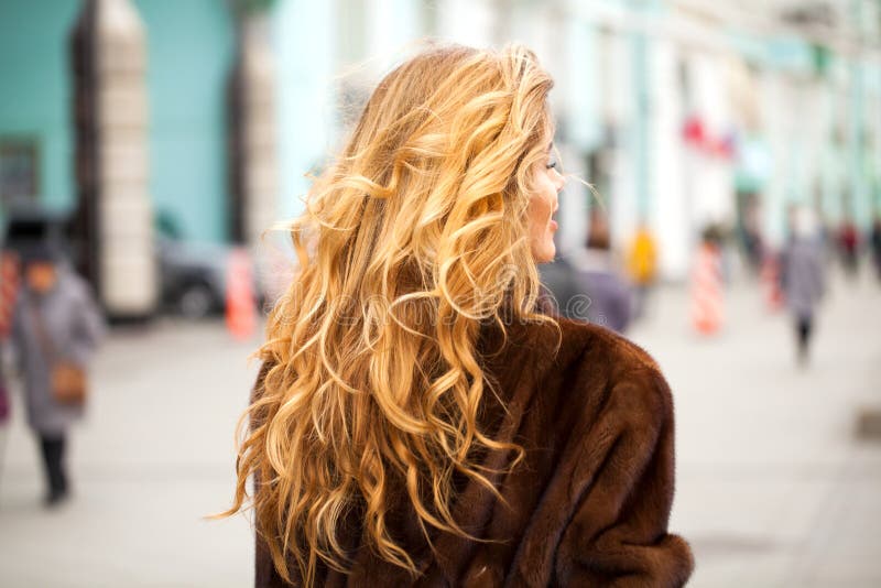 Blonde curly in fur coat walking autumn street. Healthy Wavy hair. Back view