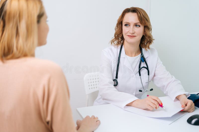 Blonde doctor wearing white lab coat sitting at desk in office and talking to young woman patient