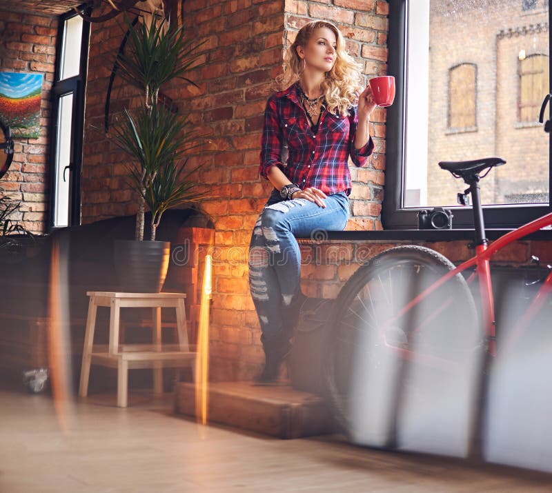 Blonde female dressed in a jeans and fleece shirt, drinks hot coffee near the window in a room with loft interior. Blonde female dressed in a jeans and fleece shirt, drinks hot coffee near the window in a room with loft interior.