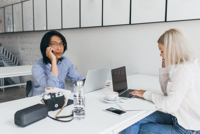 Blonde female web-developer typing on keyboard, sitting in front of asian student in glasses. Freelance programmer stock photo