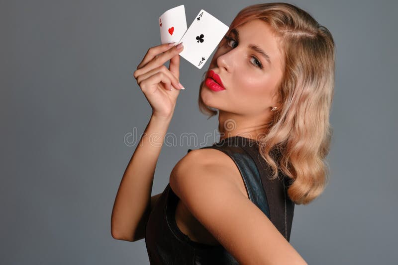 Blonde girl in black dress showing two playing cards, posing sideways against gray background. Gambling entertainment