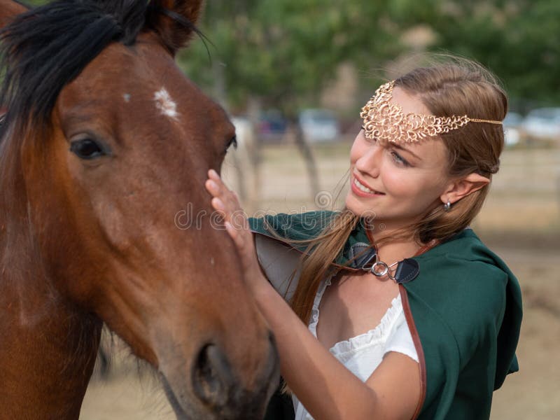 Blonde girl with blue eyes and makeup with elf in the field with a brown horse and a green cape