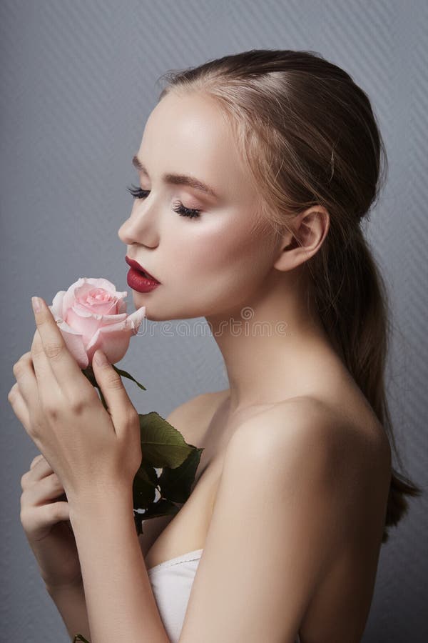 Blonde girl holding rose flowers near her face. Beauty portrait of a woman on a dark background. Perfect makeup, beautiful body