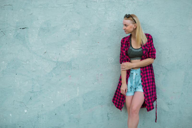 Blonde girl in light blue high-rise shorts, red plaid shirt, sneakers is posing on a blue background wall on the street