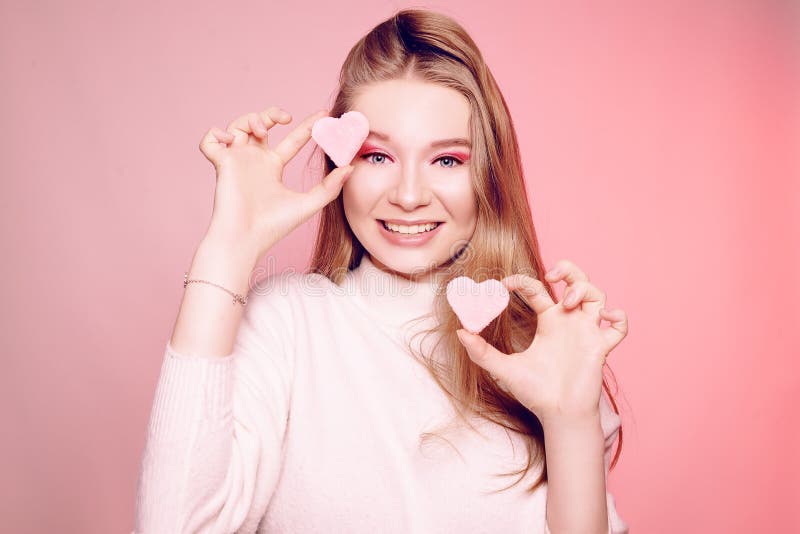 The blonde holds a sweet pink heart near her face, on a pink background on Valentine`s Day, love, discounts, Women`s Day. The gi