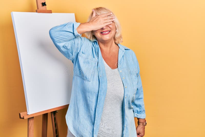 Middle age blonde woman standing by painter easel stand smiling and laughing with hand on face covering eyes for surprise. blind concept. Middle age blonde woman standing by painter easel stand smiling and laughing with hand on face covering eyes for surprise. blind concept