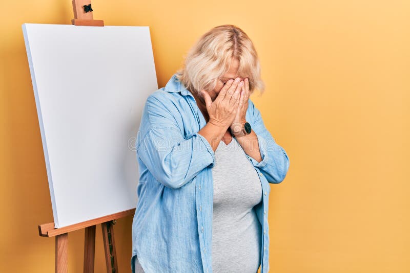 Middle age blonde woman standing by painter easel stand with sad expression covering face with hands while crying. depression concept. Middle age blonde woman standing by painter easel stand with sad expression covering face with hands while crying. depression concept