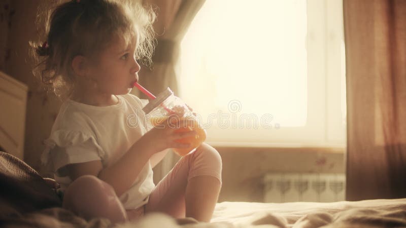 Cute blonde little girl drinks orange juice with a straw at home