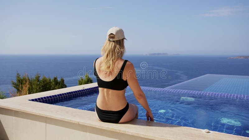 Blonde mince en maillot de bain noir et casquette beige s'asseoir au bord de la piscine et bronzer. jeune femme en bikini noir est clips vidéos