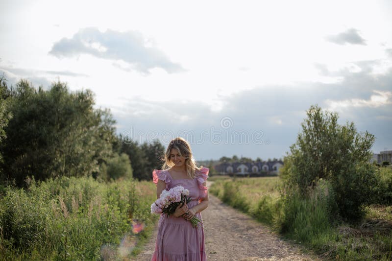Blonde romantique en robe de soleil avec bouquet de douces pivoines roses sourires marche sur la journée d'été dans la nature. photos libres de droits