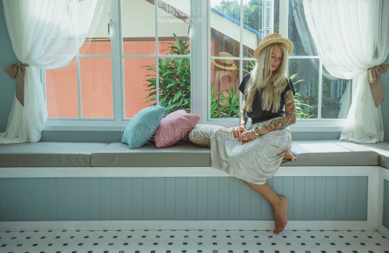 Blonde in a straw hat sits on the couch by the window. the image of a modern girl. Model with tattoos