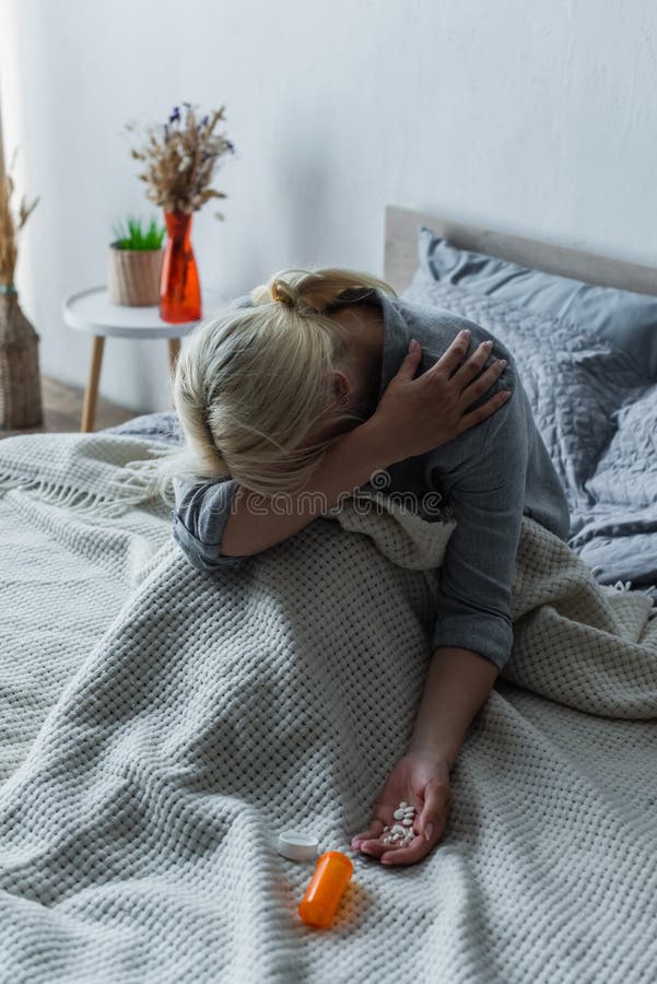 blonde woman with climax covering face while sitting in bed with painkillers in hand,stock image