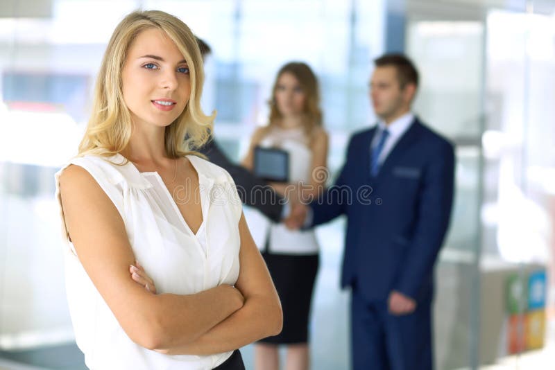 Blonde woman with touchpad computer looking at camera and smiling while business people shaking hands over background stock image Blonde woman with touchpad computer looking at camera and smiling while business people shaking hands over background stock image