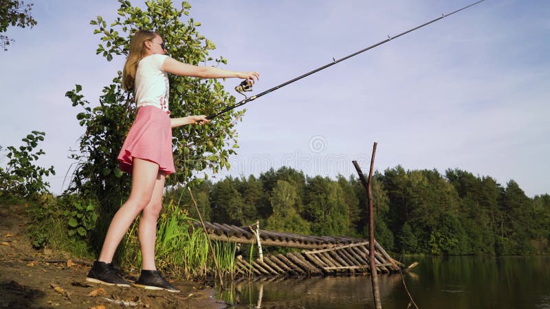 Blonde young american girl fisherman is fishing on the forest lake. woman in short dress learns to fish on big