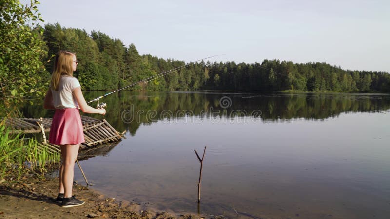 Blonde young girl fisherman is fishing on the forest lake. woman in short dress learns to fish on big river on a