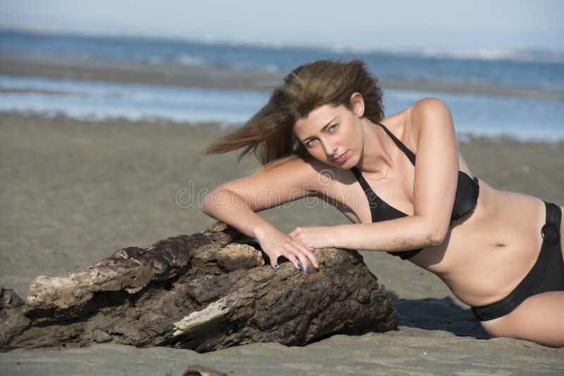 Blond woman wear black bikini lean on a tree trunks that washed up by the sea, horizontal photo. Blond woman wear black bikini lean on a tree trunks that washed up by the sea, horizontal photo
