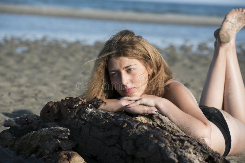 Blond woman wear black bikini lean on a tree trunks that washed up by the sea, horizontal photo. Lift her legs over her head. Blond woman wear black bikini lean on a tree trunks that washed up by the sea, horizontal photo. Lift her legs over her head