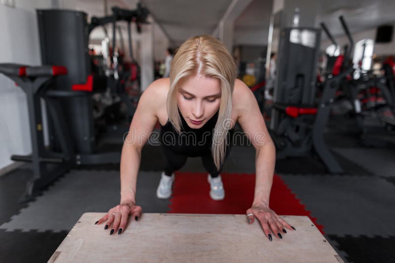 Blondinen för den unga kvinnan i sportig svart kläder utbildar i idrottshallen Flickan gör övningar för händer i konditionstudio royaltyfria foton Blondinen för den unga kvinnan i sportig svart kläder utbildar i idrottshallen Flickan gör övningar för händer i konditionstudio royaltyfria foton