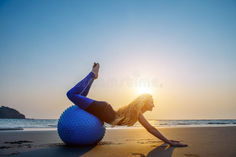 Blondinen med långt hår gör Pilates på stranden under solnedgång mot havet Ung böjlig lycklig kvinna som gör konditionövningar royaltyfri foto