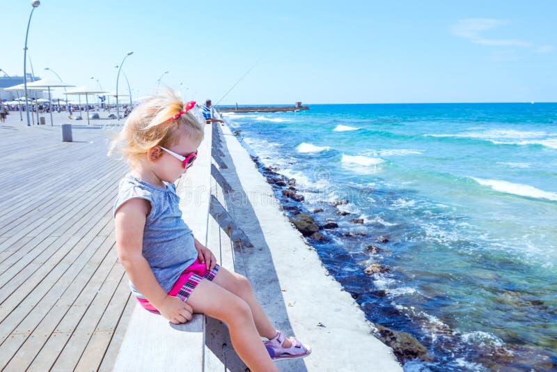 Blondy little baby girl 2-3 year old in pink sunglasses sitting alone on wooden sea railing on the seafront of Tel Aviv. Looking a