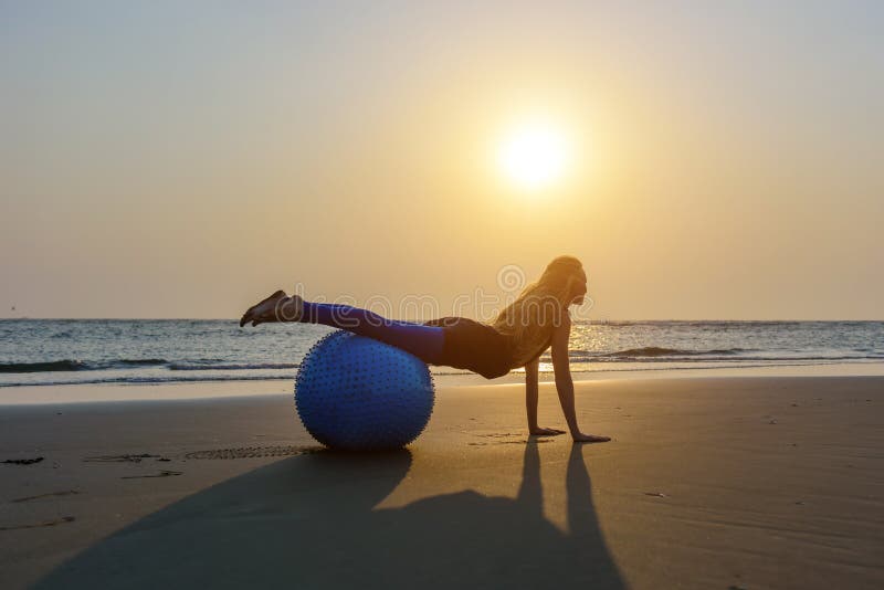 Blondynka z długie włosy robi Pilates na plaży podczas zmierzchu przeciw morzu Młoda elastyczna szczęśliwa kobieta robi sprawnośc obraz stock Blondynka z długie włosy robi Pilates na plaży podczas zmierzchu przeciw morzu Młoda elastyczna szczęśliwa kobieta robi sprawnośc obraz stock