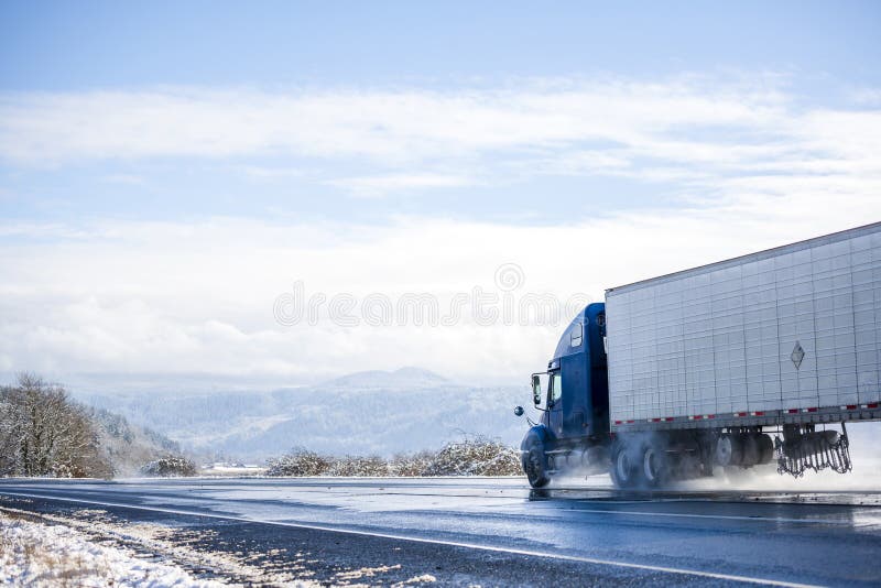 Blue big rig long haul semi truck transporting cargo in refrigerated semi trailer running on wet winter road with melting snow
