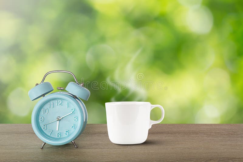 Blue classic alarm clock and hot white coffee cup with steam on vintage wooden desk on blurred green bokeh background stock images Blue classic alarm clock and hot white coffee cup with steam on vintage wooden desk on blurred green bokeh background stock images