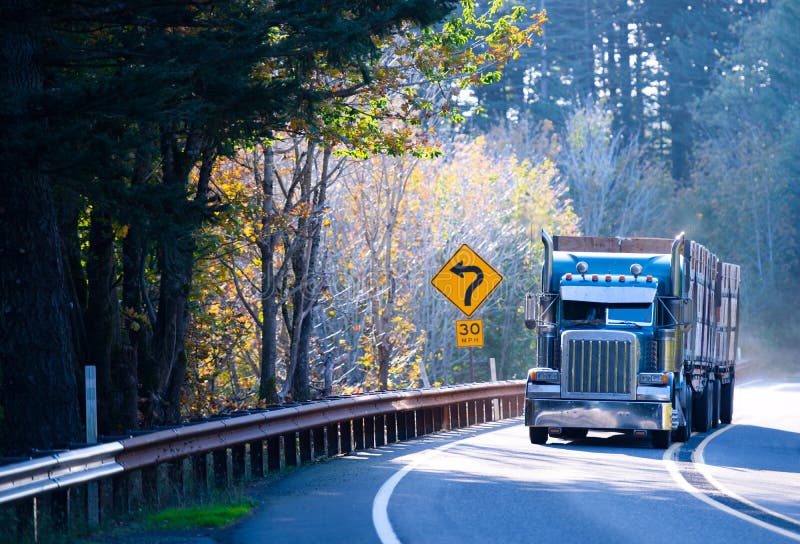 Large American professional powerful blue semi truck rig with a two trailers with high sides graft, carries loads on the winding road through the mountains, surrounded by yellowing autumn trees illuminated by the bright sun. Large American professional powerful blue semi truck rig with a two trailers with high sides graft, carries loads on the winding road through the mountains, surrounded by yellowing autumn trees illuminated by the bright sun.