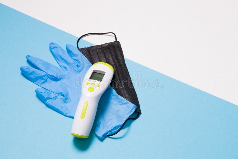 Blue disposable medical glove, black face mask, infrared thermometer for non-contact temperature measurement on a white blue background, copy space