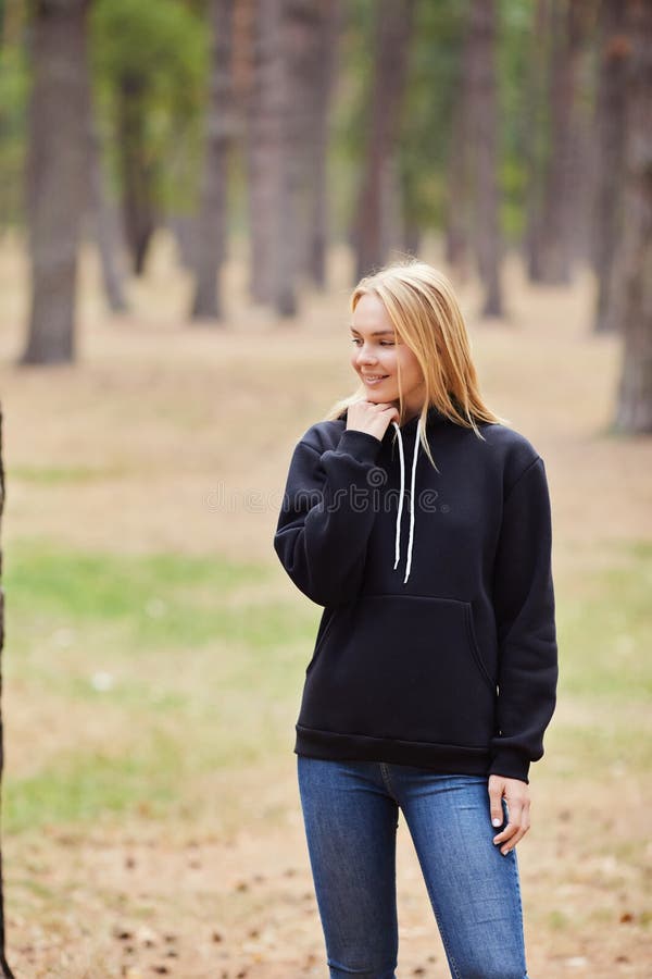 Blue-eyed blonde in a black hoodie walks in a pine forest. Portrait of a joyful young woman enjoying in autumn park