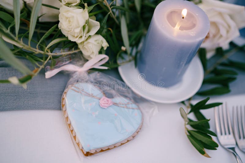 Blue heart-shaped gingerbread biscuit in a gift package lies on the table, near a burning thick wax candle in flowers