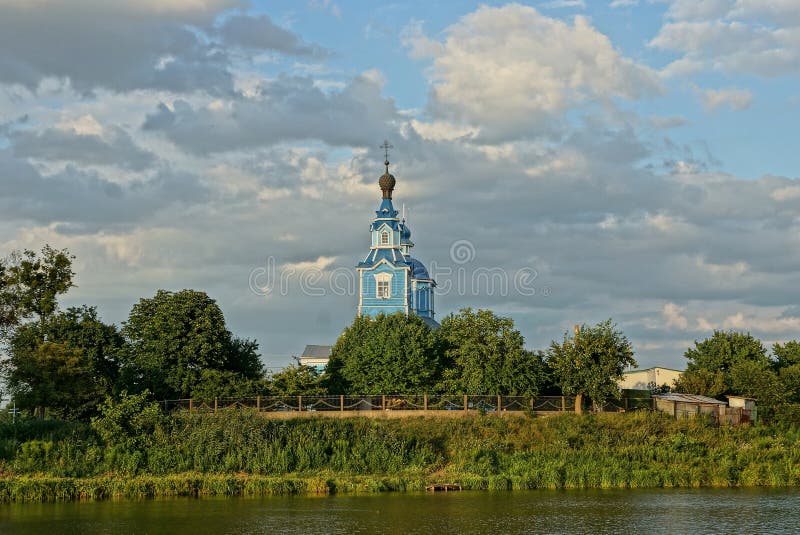 Blue old wooden church in green vegetation on the shore of the reservoir against the sky and clouds. Blue old wooden church in green trees and grass on the shore stock photography Blue old wooden church in green vegetation on the shore of the reservoir against the sky and clouds. Blue old wooden church in green trees and grass on the shore stock photography