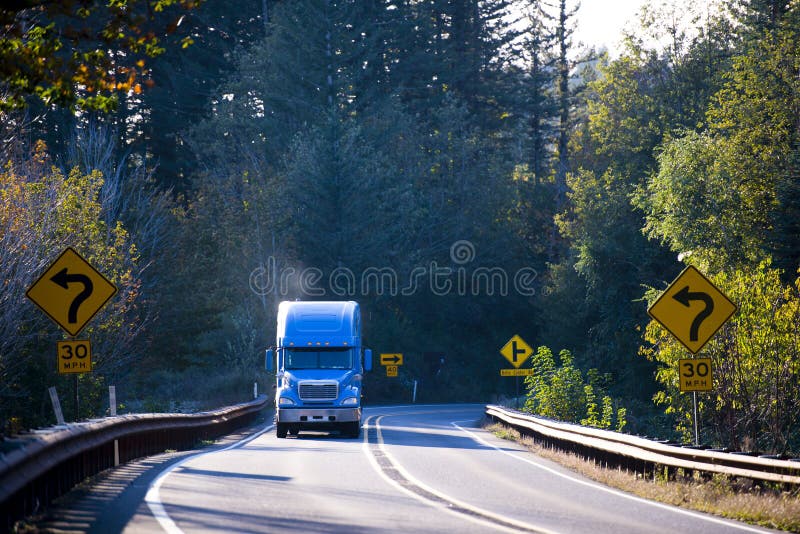 Large American professional powerful truck with smoke from the exhaust pipe, transports goods on the winding road through the mountains, surrounded by yellowing autumn trees illuminated by bright sunshine, with road signs on both sides of the road. Large American professional powerful truck with smoke from the exhaust pipe, transports goods on the winding road through the mountains, surrounded by yellowing autumn trees illuminated by bright sunshine, with road signs on both sides of the road.