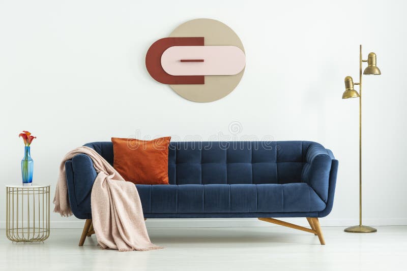 Blue sofa with cozy textiles between a golden floor lamp and a side table with flowers in a vase in a white living room interior.