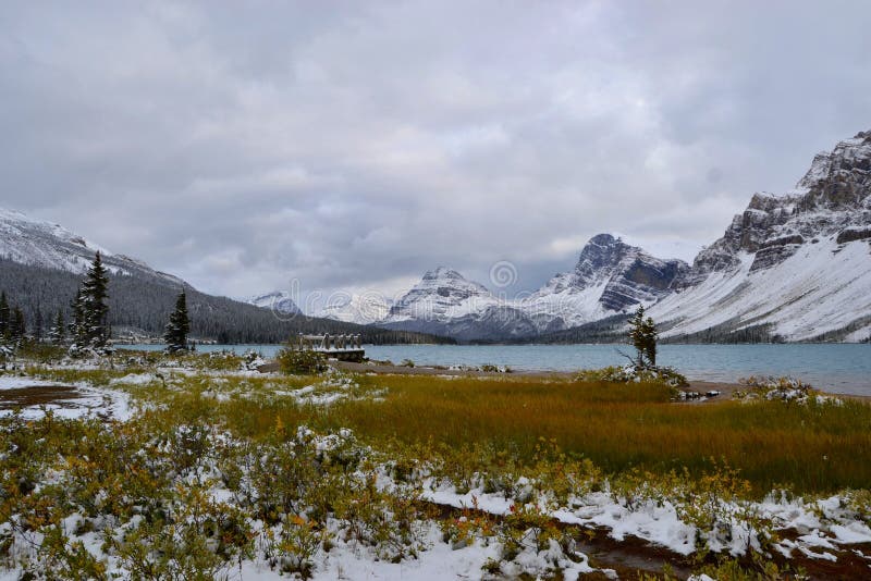 Blue water of Bow Lake, mountains covered with snow. Wooden Bridge, gray clouds, view from shore. Banff National Park, Rocky Mountains, Canada stock photos Blue water of Bow Lake, mountains covered with snow. Wooden Bridge, gray clouds, view from shore. Banff National Park, Rocky Mountains, Canada stock photos