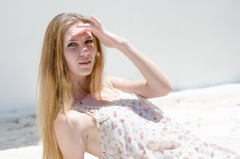 Fashion blonde wearing flower dress pose under bright sun, sitting on pavement, making shadow with her hand. Fashion blonde wearing flower dress pose under bright sun, sitting on pavement, making shadow with her hand