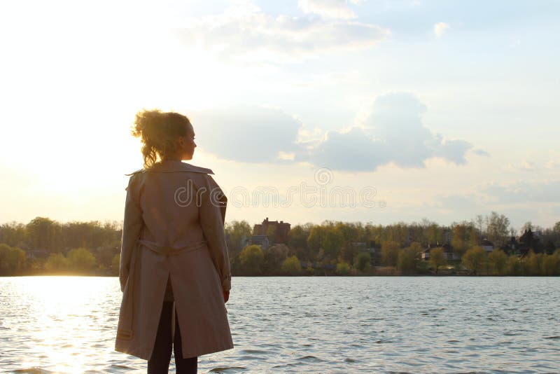 Blurry image of teenage girl wearing a beige coat and blue jeans standing near the water, overexposed shot.