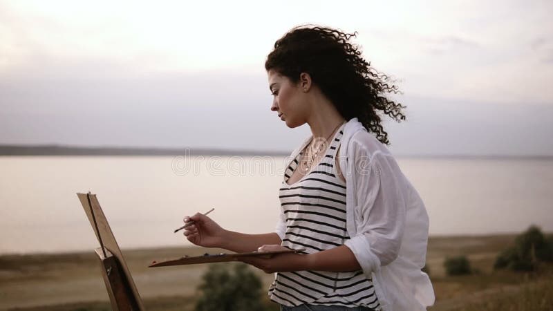 Boczny widok atrakcyjny kędzierzawy kobieta artysta pracuje na jej obrazku z sztalugą outdoors blisko jeziora Ranek, miękka część
