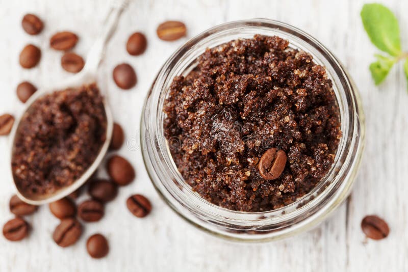 Body scrub of ground coffee, sugar and coconut oil in glass jar on white rustic table, homemade cosmetic for peeling and spa care, top view