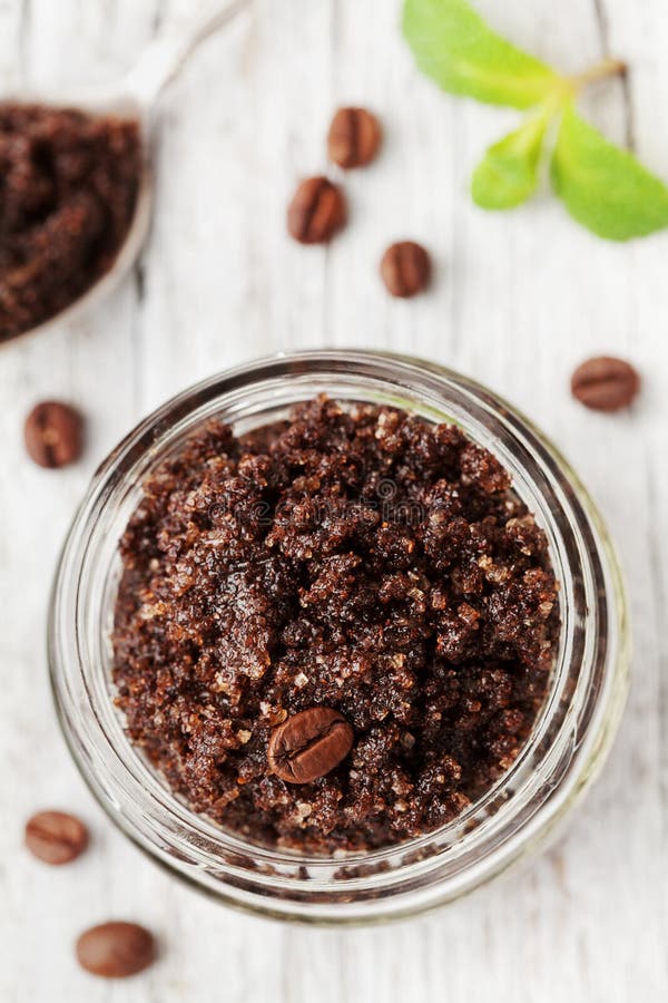 Body scrub of ground coffee, sugar and coconut oil in glass jar on white rustic table, homemade cosmetic for peeling and spa care, top view