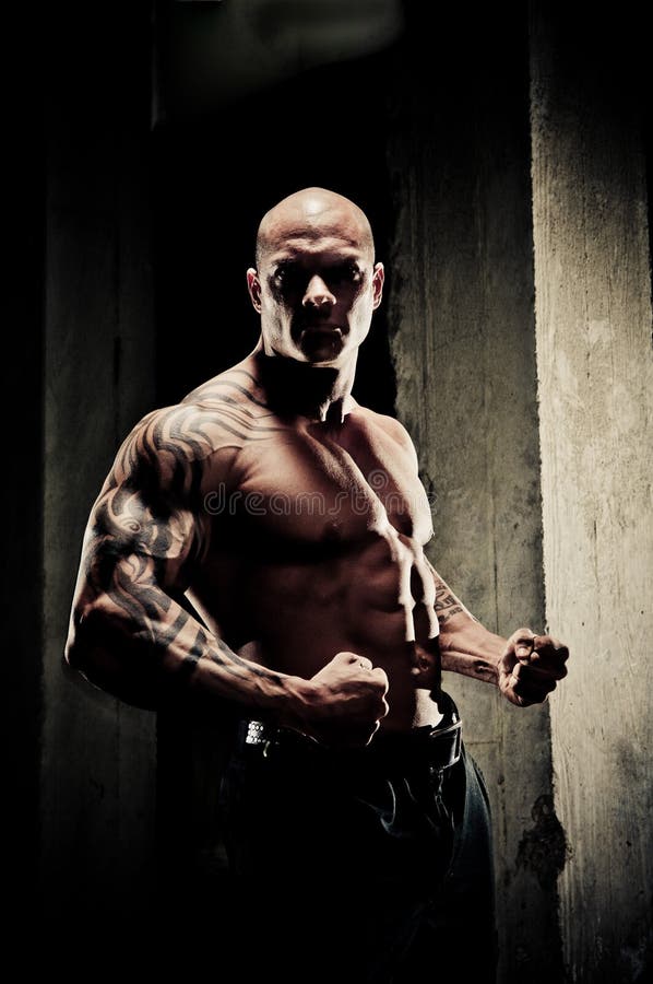 Handsome fit bodybuilder standing clenching his fists and flexing his arm muscles, dramatic lighting with highlights on musculature. Handsome fit bodybuilder standing clenching his fists and flexing his arm muscles, dramatic lighting with highlights on musculature.