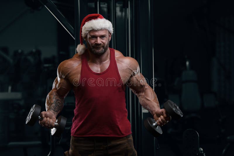 Bodybuilder in Santa Claus costume in the gym with barbell and dumbbell. tough caucasian man exercising cross fitness and bodybuilding and fitness sport concept. Trains Arms, chest and shoulders