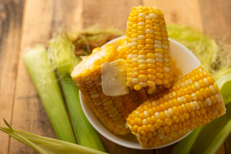 Boiled corn cob in a plate with butter, on wooden textures and green stalks of corn, tasty homemade food, for a picnic