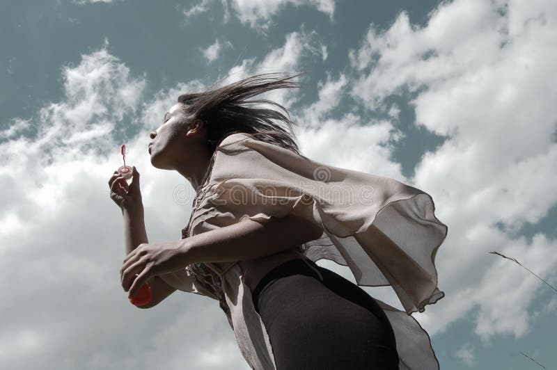 Portrait of a girl/young woman blowing soap bubbles in the wind in the summertime with the sky and clouds as background. Portrait of a girl/young woman blowing soap bubbles in the wind in the summertime with the sky and clouds as background