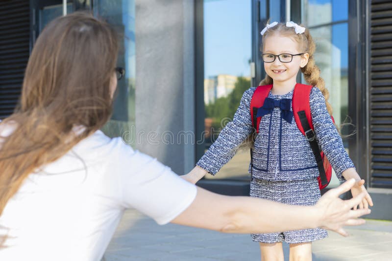 First day in the fall. back to school kids. mother lead little schoolgirl in uniform, schoolbag in the first grade. start of lessons. child wave a hand to mom. parent meeting, hugging schoolchildren. First day in the fall. back to school kids. mother lead little schoolgirl in uniform, schoolbag in the first grade. start of lessons. child wave a hand to mom. parent meeting, hugging schoolchildren